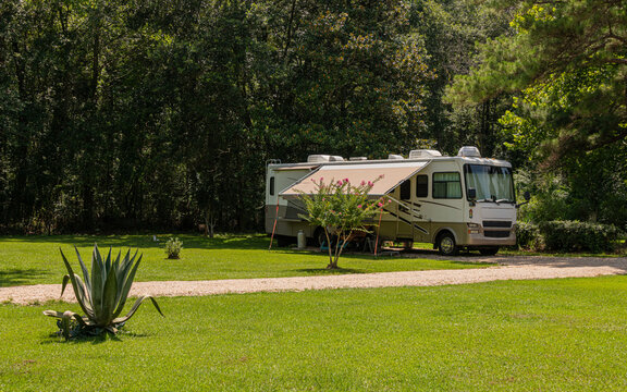 An RV is parked on a lush green lawn with trees in the background. The motorhome's awning is extended, offering shade in the sunny setting.
