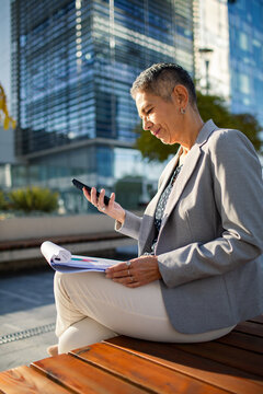 Mature businesswoman checking phone and report outside modern office building
