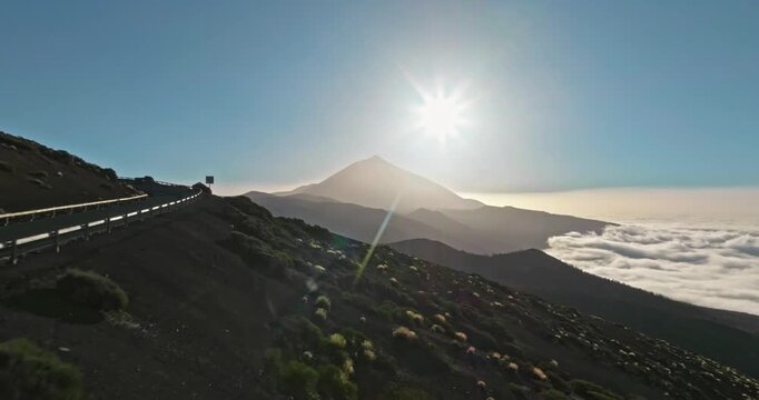 Mount Teide volcanic landscape with a winding road, fences, and a rugged hillside, showing clouds below and a bright sunburst overhead at sunset, creating a dramatic natural scene in Tenerife