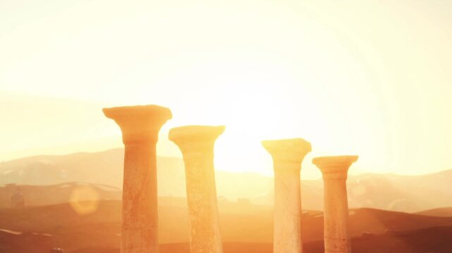 Four tall stone pillars rise proudly in a deserted area under a bright sky. The sun sets in the background, casting long shadows on the ground. The scene is quiet and relates to history.
