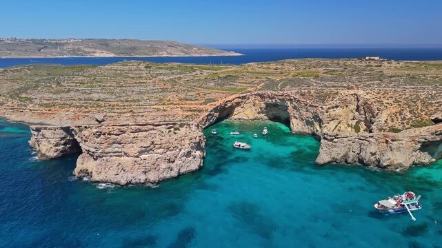 Comino, Malta, with turquoise crystal clear water, boats and swimmers along rocky cliffs and sea caves on the Mediterranean coast, captured in an aerial panorama.