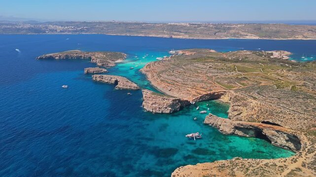 Blue Lagoon, Comino, Malta, with boats in turquoise clear water beside rocky cliffs and caves near Cominotto in the Mediterranean Sea, captured in an aerial summer view.