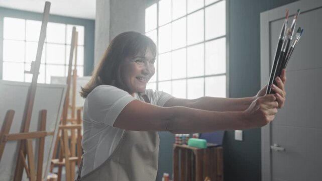 Woman smiling holds paintbrushes with both hands, arms extended toward window in art studio, facing easels and natural light; creative serenity.