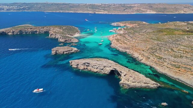 Blue Lagoon, Comino, Malta, with boats in turquoise clear water beside Cominotto cliffs and caves on the Mediterranean coast, captured by drone in sunny summer daylight.