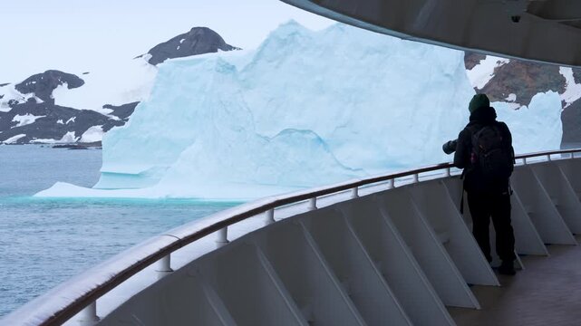 Antarctica Male Photographer on Cruise Ship Deck With Professional Lens Massive Iceberg. Exciting Adventure Travel. Nomad Black Coat, Green Hat