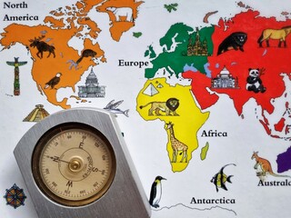 Silver metal compass placed on a colorful world map featuring animals and landmarks for travel © RJuniantoro_