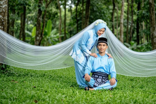 Young couple in traditional light blue attire poses for wedding photos in forest setting. Bride wearing veil, groom wears songkok cap, surrounded by green grass, trees. Romantic natural outdoor shoot.