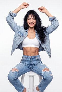 Fit young woman with a sculpted midriff throws her arms up in celebration while wearing casual double denim in a bright studio portrait.