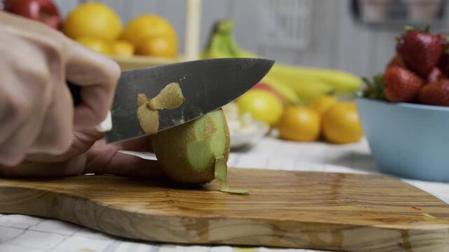 Hands carefully using sharp knife to shave skin off kiwi on cutting board - closeup
