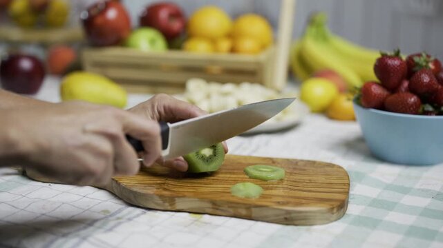 Person with sharp knife cutting slices of kiwi near other fruit in kitchen - tracking shot