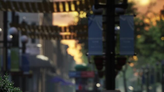People walk along a bustling street with shops and banners hanging above. The sun sets in the background, creating a warm glow. A few trees and lamp posts line the street.