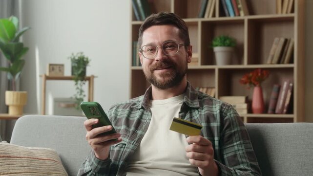 Smiling Caucasian man shopping mobile phone at home online holding credit card satisfied adult male guy couch payment buying internet purchase web money cash e-commerce buyer smartphone banking app
