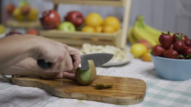 Woman shaving skin from kiwi with large sharp knife in kitchen near other fruit