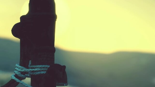 A wooden post stands firm with a rope tied to it. The sun sets in the background, casting a warm glow over the landscape. The scene shows nature at dusk.