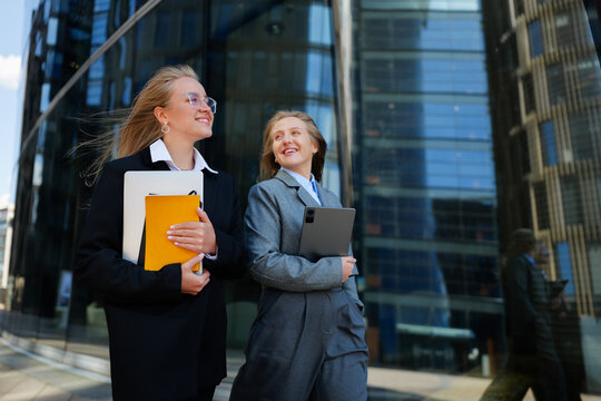 Two confident women walk side by side, enjoying a sunny day outside a modern office building while carrying digital devices and documents