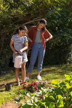 African mother watching while child watering red pink flowers in yard with pale-pink watering-can