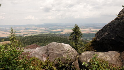 Fototapeta premium Szeroka panorama z górskiego szczytu, na którym leżą duże, zaokrąglone głazy. W dole rozciąga się gęsty zielony las oraz rozległe pola uprawne pod zachmurzonym niebem.