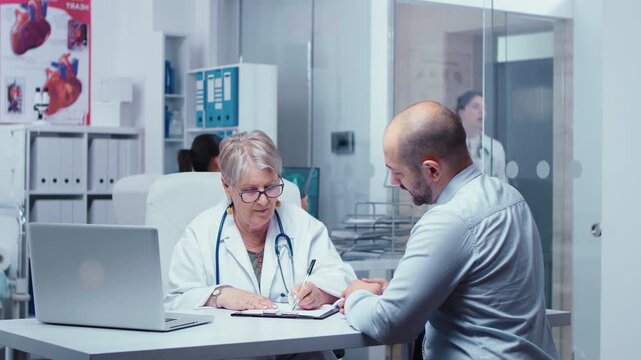 Senior woman doctor prescribes a new drug dosage to a male patient, explaining treatment medication and professional clinic expertise.