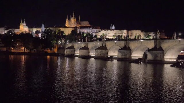 Tilt-up camera movement revealing illuminated Charles Bridge arches and Prague Castle skyline at night over the Vltava River.