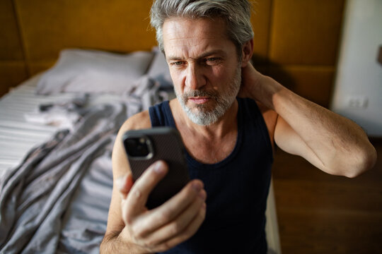 Middle-aged man with neck pain checking smartphone in bedroom