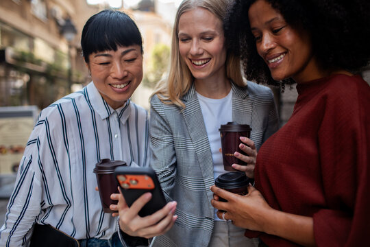 Three women friends sharing a smartphone and coffee outdoors on a city street