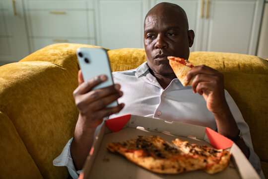 Man eating pizza while using smartphone on sofa at home