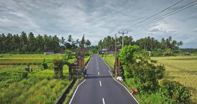 Bali's traditional Candi Bentar split gate frames a rural road passing through vibrant green rice fields and a tropical landscape under a cloudy sky, showcasing iconic Indonesian culture and scenery