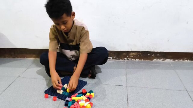 ​Little Asian boy sitting on the floor concentrating on playing with colorful educational puzzle blocks at home. 