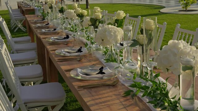 Long wooden wedding table set with white roses gold plates and glassware on a green lawn