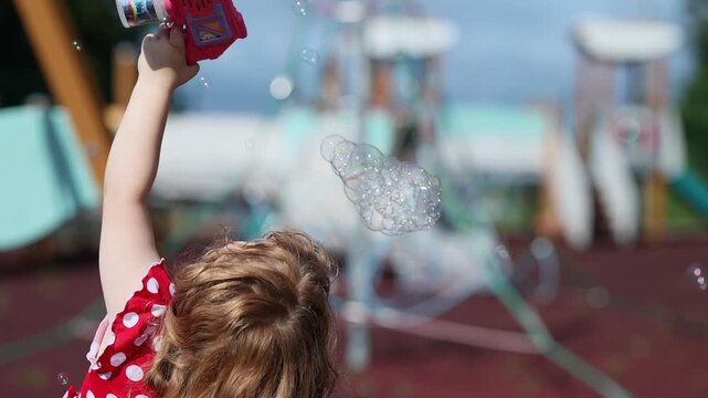 Little girl playing with bubble blower on playground. Caucasian child in red polkadot dress making soap bubbles. Happy childhood activity and outdoor summer game.