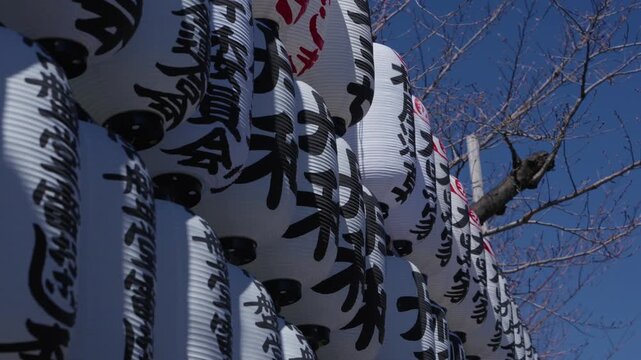 Rows of white paper lanterns with black kanji calligraphy against a blue sky at a Japanese shrine