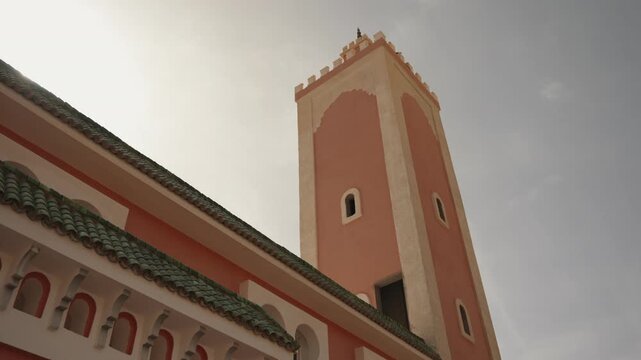 Pink plaster minaret and green tiled roof of a Morocco mosque under a pale hazy sky