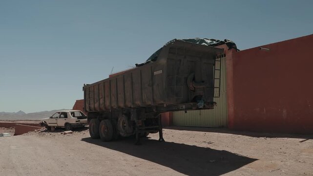 Rusty tipper trailer parked beside a red wall with an abandoned car on sand in Morocco