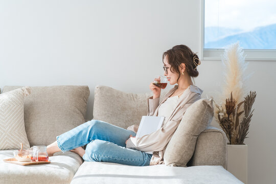 A stylish Asian woman sitting on the living room sofa, reading a magazine during teatime.