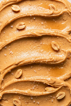 Macro close up texture of creamy peanut butter spread with sea salt crystals and roasted peanut halves background