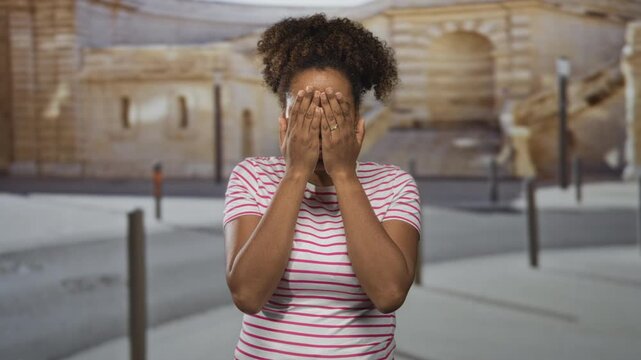 Woman sticks tongue out and frames her face with both hands while standing near bollards and a parked car on an urban street; playful mischief.