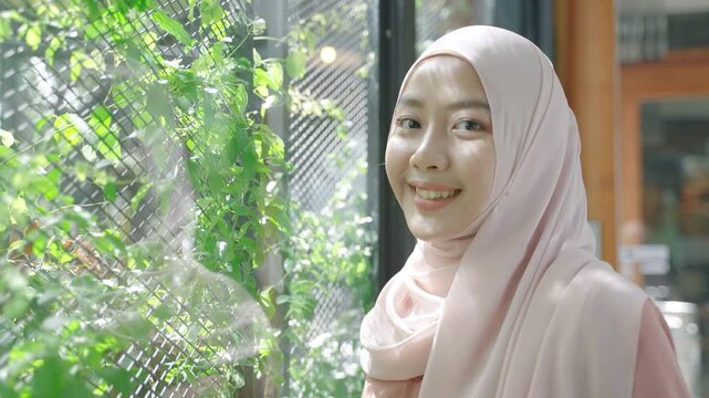 Portrait of a smiling young Muslim woman wearing a light-colored hijab beside a green vertical garden. Natural sunlight, modest fashion, positive lifestyle, and multicultural beauty concept.
