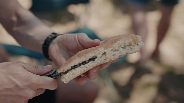 Hands cutting a crusty bread roll open with a pocket knife at an outdoor gathering
