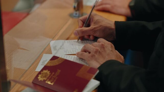 Hands signing an arrival immigration form beside a passport and travel papers at a border desk