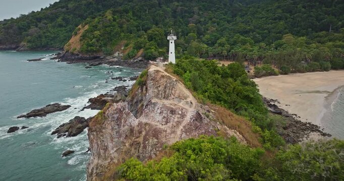 Waterfall Bay Lighthouse standing on a rocky cliff overlooking the turquoise sea and a secluded sandy beach surrounded by lush green tropical forest in Koh Lanta, Thailand. Aerial zoom out panorama