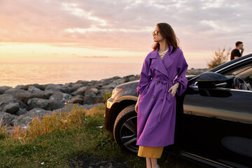 Fototapeta premium A woman in a purple coat stands beside a dark car at sunset, gazing toward the horizon with a confident pose as warm light reflects off rocks and calm water nearby.