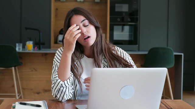 A distressed young woman experiences a terrible headache and stress while trying to concentrate on her laptop at a wooden table in her modern home office setup.