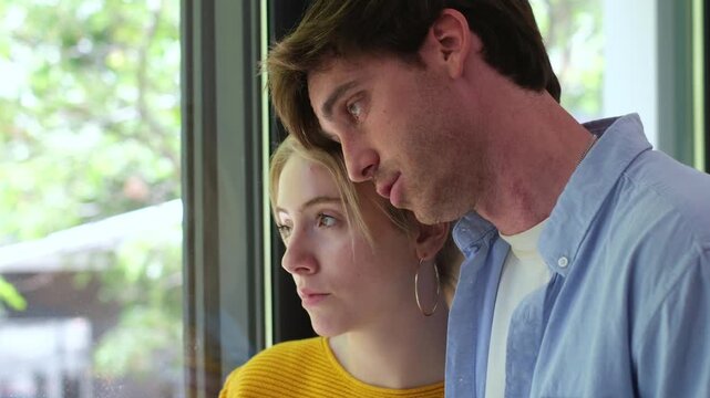 Pensive young couple embracing and looking out the window, showing sadness, concern, and contemplation as they face a difficult situation together, finding comfort in their relationship