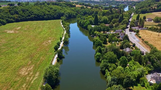 Drone view flying over calm Mayenne river surrounded by lush green trees and agricultural fields in France. Aerial forward