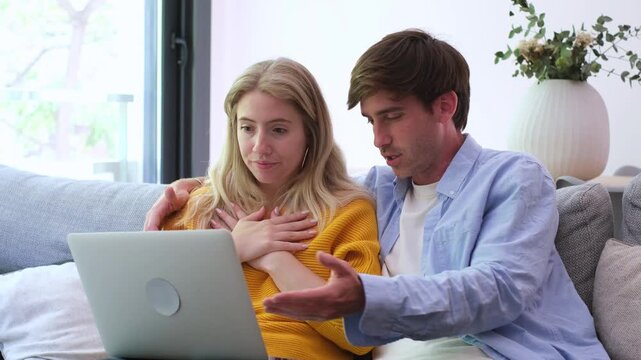 A young couple sits on their couch at home, reacting with distress and concern as they discover a significant problem or bad news while looking at their laptop screen together.