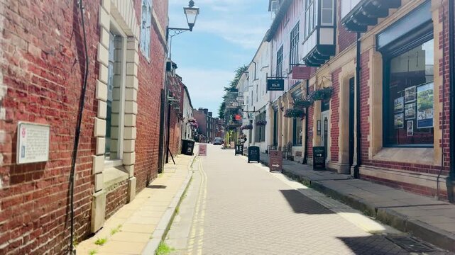 View along historic narrow street in Winchester city centre, lined with brick buildings, independent shops, traditional architecture, and quiet pedestrian walkway during sunny summer day