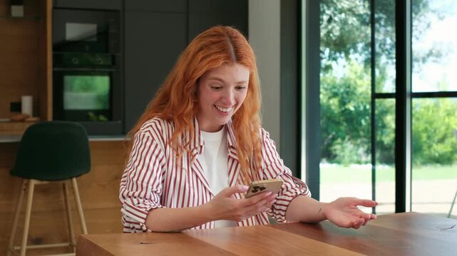 Happy young woman with red hair looking at her smartphone and celebrating success. A victory. Or good news while sitting at a table in a modern kitchen. She makes a winning gesture. Feeling ecstatic
