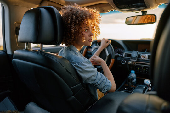 Young woman driver sits in a car, smiling gently, hands on the steering wheel. Bright natural light fills the modern interior, conveying a casual confident lifestyle and everyday travel.