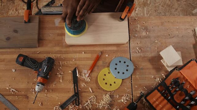 Top view of craftsman sanding wooden board with orbital sander on workbench surrounded by woodworking tools, drill, clamps and wood shavings
