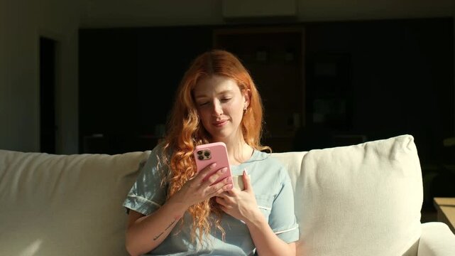 Beautiful young woman with long red hair sitting on the couch in the morning sunlight, smiling while scrolling on her smartphone and holding a cup of coffee, enjoying a relaxing moment at home